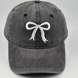 Puede incluir: Una gorra de béisbol gris carbón con un acabado desgastado y lavado. El panel frontal presenta un diseño de lazo bordado en blanco centrado sobre la visera. La gorra incluye una visera curva y ojales negros en la corona para ventilación.