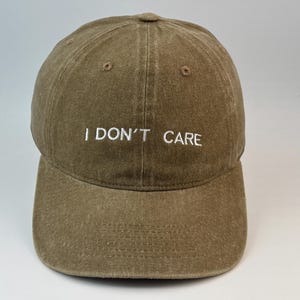 May include: A brown baseball cap with a washed-out, vintage-style finish. The front of the cap features the text I DON'T CARE embroidered in white capital letters. The hat has a structured crown, a curved brim, and matching brown stitching.