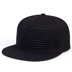 May include: A solid black baseball cap featuring a structured crown and a flat brim. The front panel displays a tonal, embroidered American flag design, where the stars and stripes are rendered in the same black thread as the cap fabric.