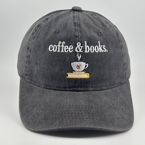 May include: A charcoal grey baseball cap featuring white embroidered text that reads coffee & books. Below the text is an embroidered graphic of a white coffee cup with steam rising, resting on a yellow book with a white page edge detail.