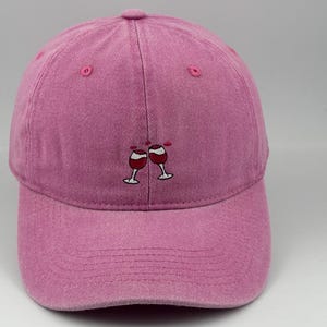 Pode incluir: Um boné de beisebol rosa com um bordado de duas taças de vinho a brindar. O boné tem uma copa estruturada, uma aba curva e ilhós rosa a condizer para ventilação, centrado com o pequeno desenho de taças de vinho em vermelho e branco.