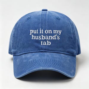 Peut inclure: Une casquette de baseball bleue avec une finition délavée et usée. L'avant de la casquette présente un texte brodé en blanc qui indique : "put it on my husband's tab". La casquette possède une visière courbée, un bouton sur le dessus.