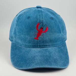 Può includere: Un cappellino da baseball blu con un ricamo raffigurante un'aragosta rossa al centro del pannello frontale. Il cappellino presenta una corona strutturata, una visiera curva con cuciture a vista e un bottone sulla parte superiore. Il tessuto è sbiadito.