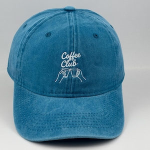 Pode incluir: Um boné de beisebol azul com texto bordado em branco que diz Coffee Club acima de um gráfico de linhas de duas mãos segurando canecas de café. O boné tem uma aba curva, um botão no topo e ilhós costurados para ventilação na coroa.