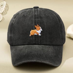 Peut inclure: Une casquette de baseball noir anthracite avec une texture délavée. Le centre avant présente une petite broderie détaillée d'un chien Corgi marron et blanc en position de course. La casquette a une visière incurvée et un bouton au sommet.