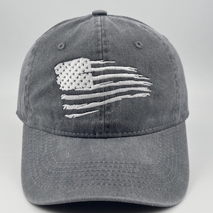 May include: A charcoal grey baseball cap featuring an embroidered white design of a distressed American flag. The cap has a six-panel construction with a curved brim and a button on the top centre, showcasing a weathered fabric texture.