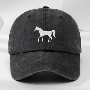May include: A charcoal black baseball cap with a washed, distressed texture. The front panel features a white embroidered silhouette of a horse in a standing position. The cap has a curved brim and a structured crown with stitched eyelets.
