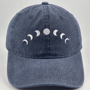 May include: A dark blue denim baseball cap featuring an embroidered white moon phase cycle across the front panels. The design displays a series of crescent moons transitioning into a full moon, arranged in a curved arc pattern on the fabric.