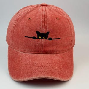 Può includere: Un cappellino da baseball rosso con una finitura slavata e usurata. La parte anteriore presenta la sagoma ricamata di un gatto nero con occhi gialli, che sembra spuntare sopra una linea nera orizzontale che attraversa il centro dei pannelli.