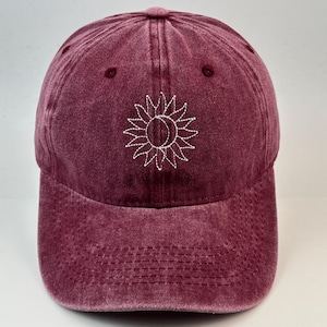 Könnte beinhalten: Eine kastanienbraune Baseballkappe mit verwaschener Textur. Auf der Vorderseite befindet sich eine weiße Stickerei, die eine Sonne mit einer Mondsichel in der Mitte darstellt. Die Kappe hat einen gebogenen Schirm mit sichtbaren Nähten und Ösen.