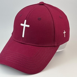 Puede incluir: Una gorra de béisbol de color burdeos intenso con una cruz blanca bordada centrada en el panel frontal y una cruz blanca más pequeña bordada en el panel lateral. La gorra tiene una corona estructurada, visera curva con costuras y un botón.