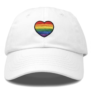 May include: A white baseball cap featuring an embroidered heart patch on the front centre. The heart is composed of horizontal stripes in red, orange, yellow, green, blue, and purple, outlined in black thread against the white fabric of the cap.