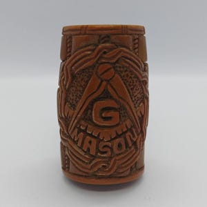 May include: A brown, cylindrical object with carved Masonic symbols. The design includes a compass, the letter "G", and the word "MASON" in a decorative font. The object is likely made of wood or a similar material.