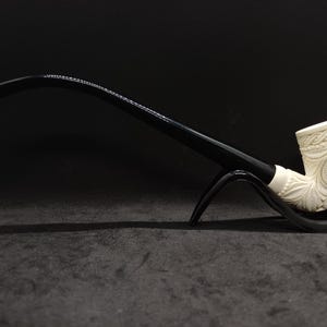 May include: A carved ivory-colored tobacco pipe with a black stem and mouthpiece. The bowl features floral and geometric designs. The pipe rests on a black stand against a dark background.