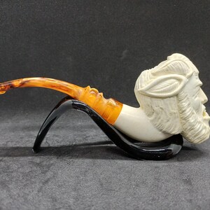 Peut inclure: Une pipe à tabac sculptée représentant un visage barbu avec une couronne de laurier. Le foyer est de couleur crème, le tuyau orange, et elle est présentée sur un support noir. La tige est incurvée.