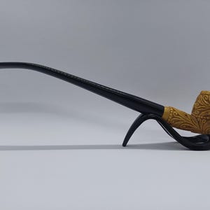 May include: A carved wooden tobacco pipe with a black stem and a decorative bowl. The bowl features intricate floral carvings. The pipe rests on a black stand, showcasing its elegant design. The pipe is a light brown color.