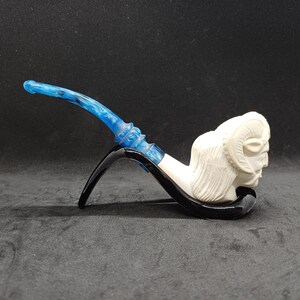 Peut inclure: Une pipe à tabac avec un foyer blanc en forme de tête de bélier. La tige est bleu et blanc marbré, et la pipe a une base noire. La pipe est posée sur un fond sombre.