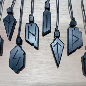 Può includere: Una collezione di nove ciondoli runici in legno nero, ciascuno sospeso a un cordino nero. I ciondoli presentano simboli runici scolpiti in varie forme geometriche. I ciondoli sono esposti su uno sfondo chiaro.