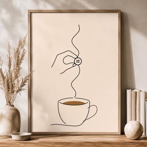 May include: A framed wall art print featuring a minimalist black line drawing on a beige background. The illustration depicts a hand holding a four-hole button above a coffee cup, with a wavy line connecting the button to the brown liquid inside.