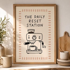 Könnte beinhalten: Ein gerahmter Kunstdruck auf cremefarbenem Hintergrund mit einer schwarzen Strichzeichnung einer Espressomaschine und einer Kaffeetasse. Der Text THE DAILY RESET STATION steht in schwarzen Buchstaben, umrahmt von kleinen rosa Strichen.