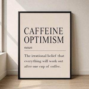 Op de afbeelding: Een ingelijste kunstprint met een definitie in woordenboekstijl. De tekst luidt: CAFFEINE OPTIMISM noun The irrational belief that everything will work out after one cup of coffee. De print heeft zwarte tekst op een lichtbeige achtergrond.