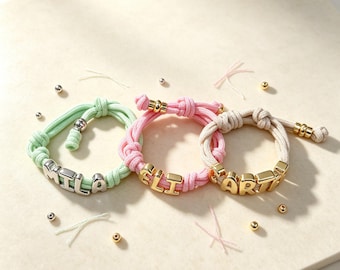 May include: Three adjustable cord bracelets in light green, pink, and beige. Each bracelet features knotted cord ends and metallic letter beads spelling out the names MILA, ELI, and ARTI, accented with small metallic spacer beads along the cord.