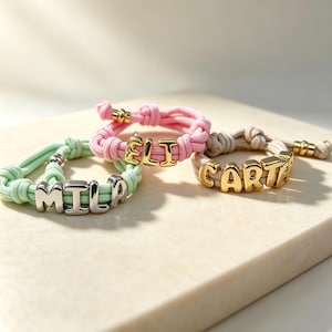 Può includere: Tre braccialetti in corda regolabili con perline a lettera metalliche. Un braccialetto verde chiaro riporta la scritta MILA in argento, uno rosa riporta ELI in oro e uno beige riporta CARTA in oro. Ogni braccialetto ha estremità annodate e dettagli metallici.