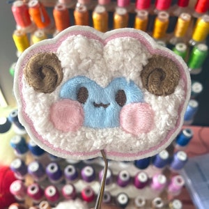 May include: A fluffy white sheep with brown horns and pink cheeks, embroidered on a pink fabric patch. The patch is being held by a pair of tweezers.