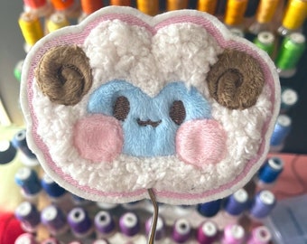 May include: A fluffy white sheep with brown horns and pink cheeks, embroidered on a pink fabric patch. The patch is held by a pair of tweezers.