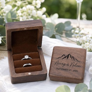 May include: A dark brown wooden ring box with a hinged lid, featuring a mountain graphic, the names Avery & Nolan, and the date September 21, 2025, engraved on the top. The interior is lined with brown velvet and holds two silver rings.