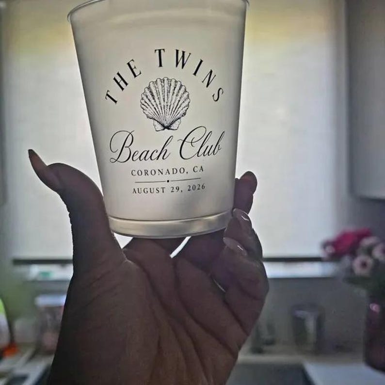 May include: A frosted plastic cup featuring a black graphic of a seashell centered above the text THE TWINS Beach Club, CORONADO, CA, and the date AUGUST 29, 2026. The cup has a smooth, translucent surface and a tapered cylindrical shape.