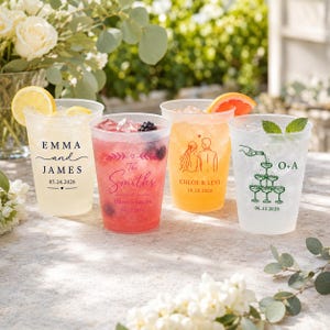 May include: Four clear plastic cups filled with ice and beverages, each featuring custom printed designs. The cups display names like Emma and James, The Smiths, Chloe and Levi, and initials O+A, along with wedding dates and graphic illustrations.
