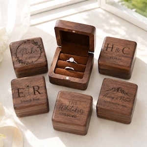 Puede incluir: Cinco cajas cuadradas para anillos de madera oscura con tapas abatibles e interiores de terciopelo marrón. Una caja está abierta, mostrando dos anillos de plata. Cada tapa presenta grabados láser únicos, incluyendo nombres como Parker, H&C, E&R, The Whitakers y Avery & Nolan.