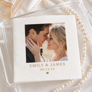 May include: A square white paper napkin featuring a centered photograph of a couple embracing. Below the image, the text EMILY & JAMES is printed in gold, followed by the date 08.24.24 and a small gold heart icon centered at the bottom edge.