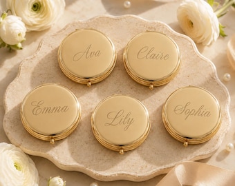 May include: Five round gold-toned metal compact mirrors, each featuring a unique name engraved in a cursive script. The names displayed on the polished surfaces are Ava, Claire, Emma, Lily, and Sophia. Each compact has a small hinge and clasp.