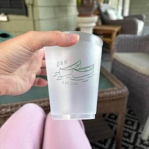 May include: A translucent, frosted plastic cup featuring a dark green graphic of two canoes on water. Above the canoes, the text reads O & N, and below the canoes, the date 7.23.2026 is printed in a simple, dark green sans-serif font.