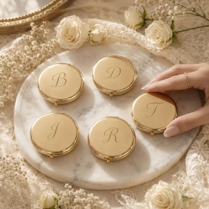 May include: Five round gold-toned metal compact mirrors, each engraved with a single cursive letter B, D, J, R, or T. The polished metallic surfaces reflect light, and each compact features a small hinge and clasp mechanism on the side.