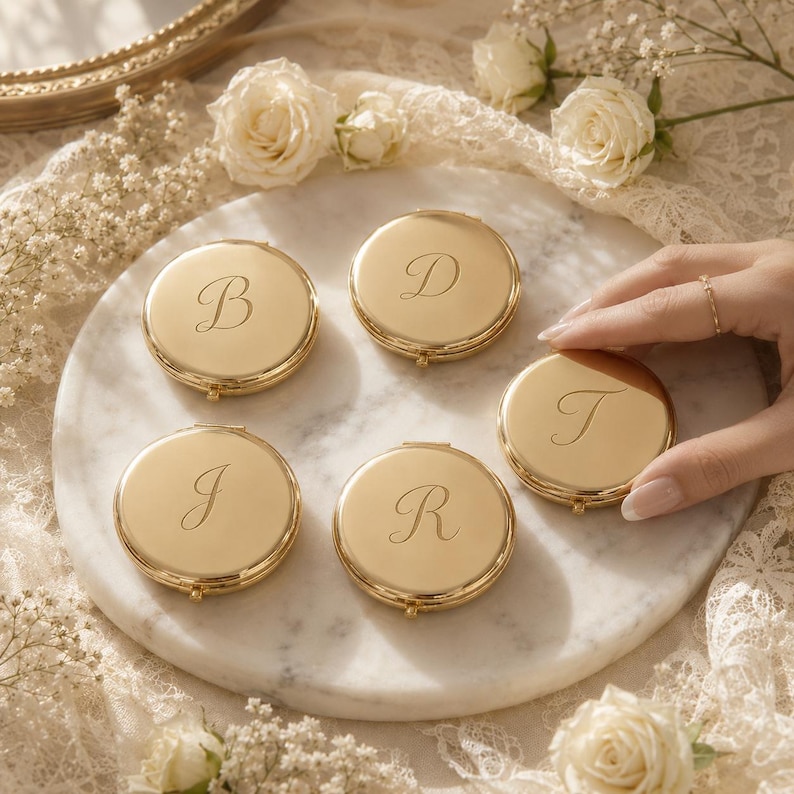 May include: Five round gold-toned metal compact mirrors, each engraved with a single cursive letter B, D, J, R, or T. The polished metallic surfaces reflect light, and each compact features a small hinge and clasp mechanism on the side.