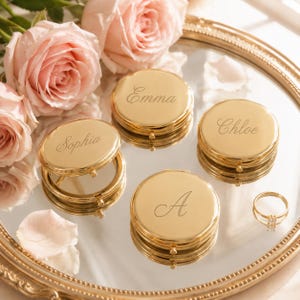May include: Four round gold-toned metal compact mirrors with engraved names Emma, Sophia, Chloe, and the letter A, arranged on a circular mirrored tray alongside a thin gold ring and several soft pink rose blossoms with scattered petals.