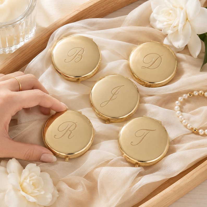 May include: Five round gold-toned metal compact mirrors, each featuring a single engraved script letter. The letters B, D, J, R, and T are centred on the polished surfaces. These small, circular cases have a smooth, reflective finish.