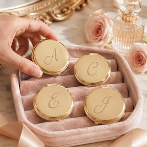 May include: Four round gold-toned metal compact mirrors, each engraved with a single cursive letter A, C, E, or J. The compacts are arranged inside a soft, dusty pink velvet jewellery tray, showcasing a polished, reflective surface finish.