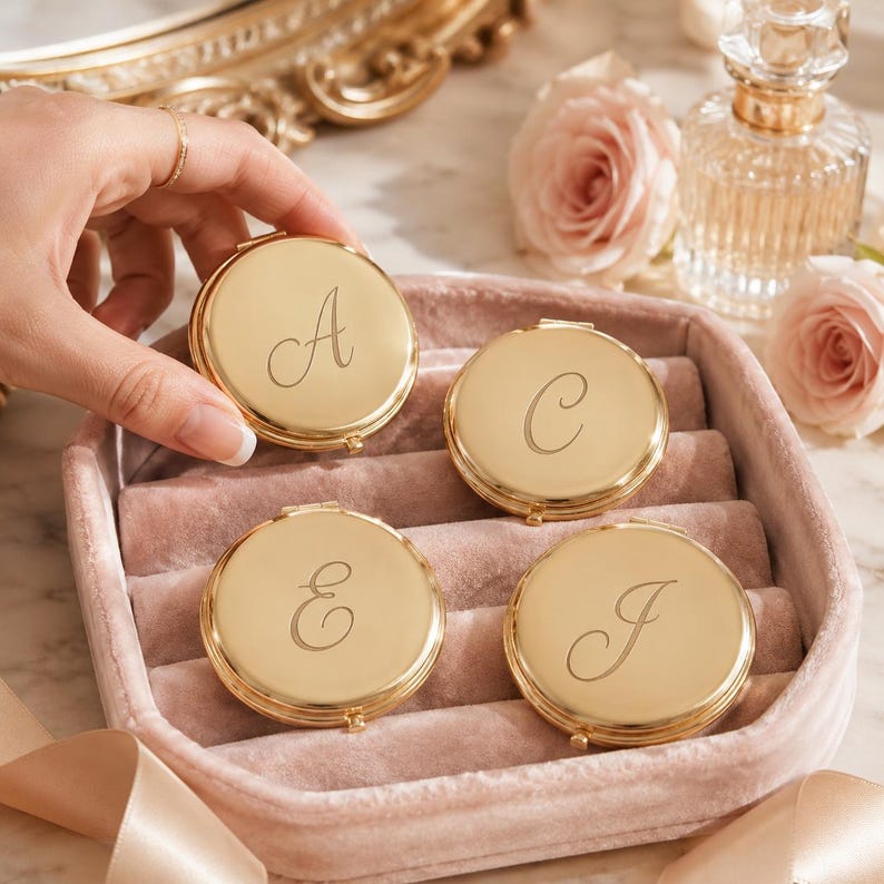 May include: Four round gold-toned metal compact mirrors, each engraved with a single cursive letter A, C, E, or J. The compacts are arranged inside a soft, dusty pink velvet jewellery tray, showcasing a polished, reflective surface finish.