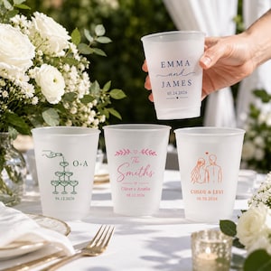 May include: Four frosted plastic cups featuring custom wedding designs. One cup held in a hand displays "EMMA and JAMES 05.24.2026". Others show a champagne tower with "O+A 06.12.2026", "The Smiths" with "Oliver + Amelia 08.12.2026", and a couple with "CHLOE & LEVI 10.10.2026".