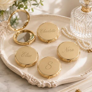May include: A collection of round, gold-toned metal compact mirrors. One mirror is open, revealing two reflective surfaces. The other three closed compacts are engraved with the names Charlotte, Elise, and Nora, and the letter S in script font.