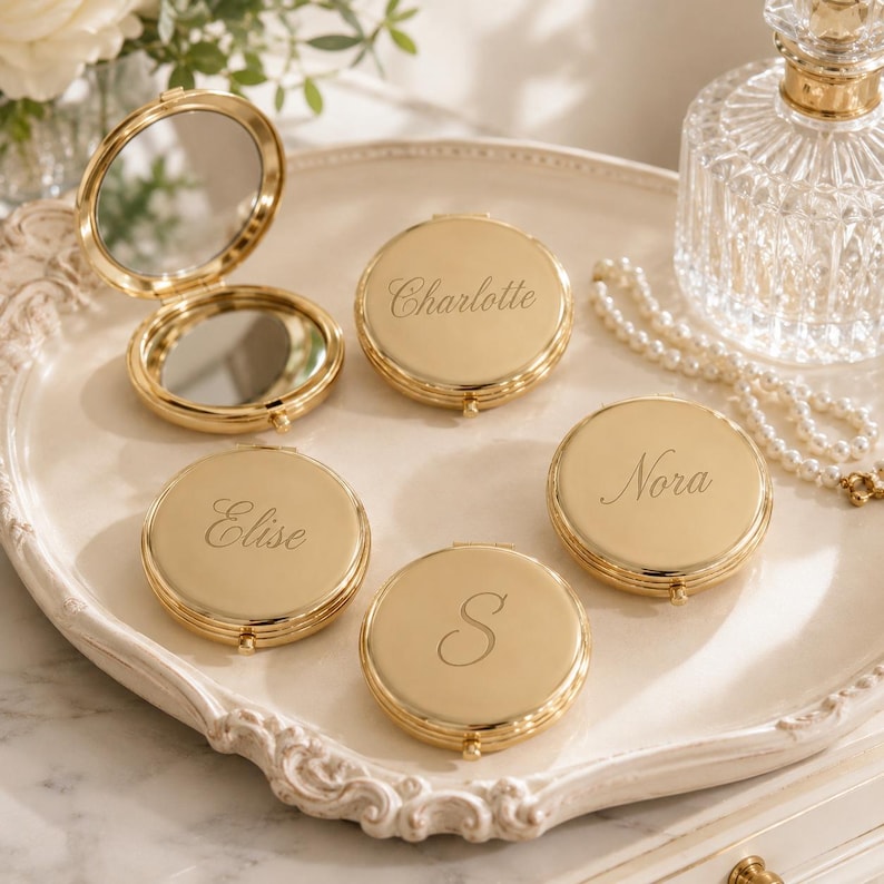 May include: A collection of round, gold-toned metal compact mirrors. One mirror is open, revealing two reflective surfaces. The other three closed compacts are engraved with the names Charlotte, Elise, and Nora, and the letter S in script font.