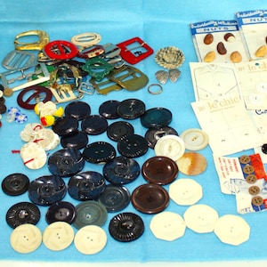 May include: A large collection of assorted sewing buttons in various sizes, shapes, and colors, including loose buttons in a pile, several metal buckles, and multiple retail cards featuring buttons and fasteners with various brand names and text.