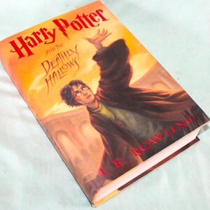 May include: A hardcover book titled "Harry Potter and the Deathly Hallows" by J.K. Rowling. The cover features an illustration of Harry Potter with his arm raised, set against a warm, orange and yellow background.
