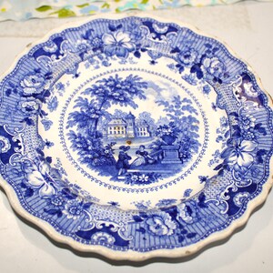 May include: A blue and white ceramic plate with a scalloped edge. The plate features a detailed landscape scene in the center, surrounded by floral and geometric patterns. The color palette is primarily blue and white, with a slightly aged appearance.