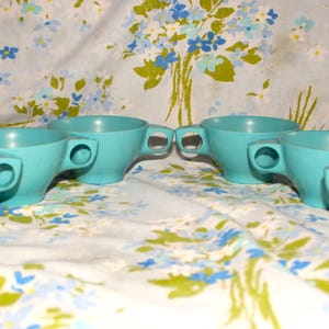 May include: Six turquoise-colored teacups with rounded handles are arranged in a semi-circle. The cups are made of a solid, smooth material. The background is a floral patterned fabric with blue and white flowers.