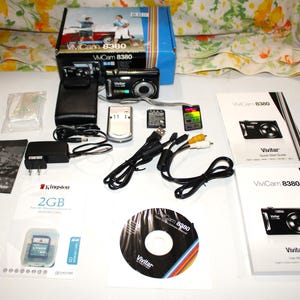 May include: A Vivitar Vivicam 8380 digital camera kit, including the camera, a case, charger, cables, memory cards, and manuals. The camera is black with silver accents. The kit includes a 2GB Kingston memory card and a Vivitar CD.